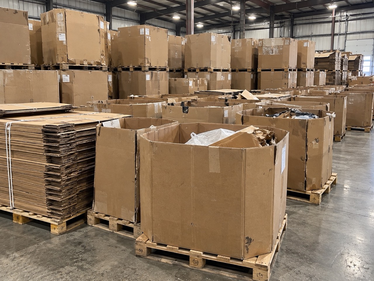 Rows of used gaylord boxes stacked two high on pallets in a warehouse aisle, with banded stacks of flattened corrugated alongside