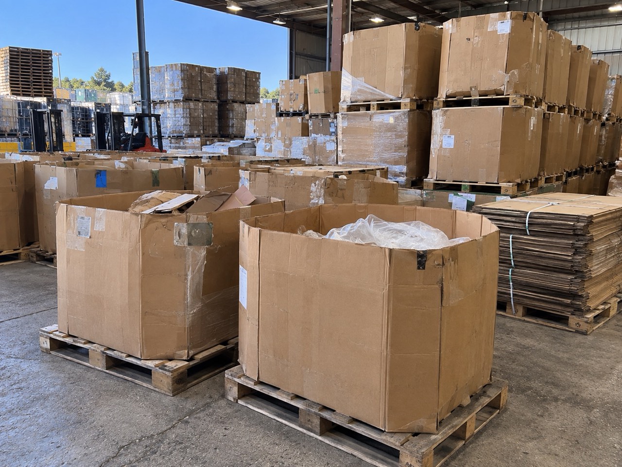Open warehouse dock with used bulk boxes on pallets in the foreground and a forklift and pallet stacks in the yard beyond